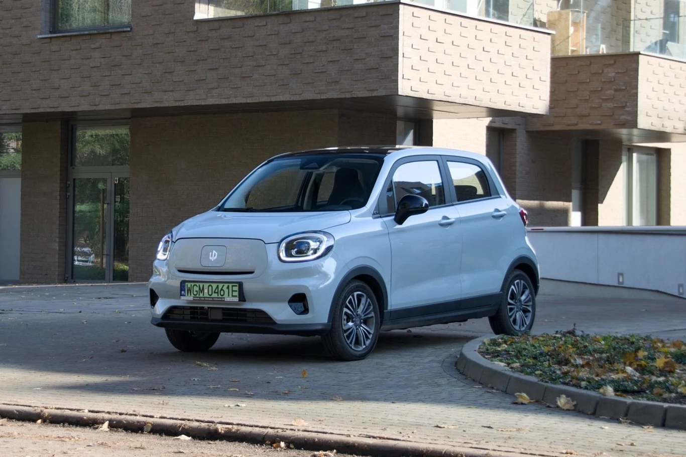 Jasnoszary samochód elektryczny zaparkowany na podjeździe przed nowoczesnym budynkiem z dużymi oknami. Otoczenie jest uporządkowane, widoczny fragment rabaty z liśćmi.