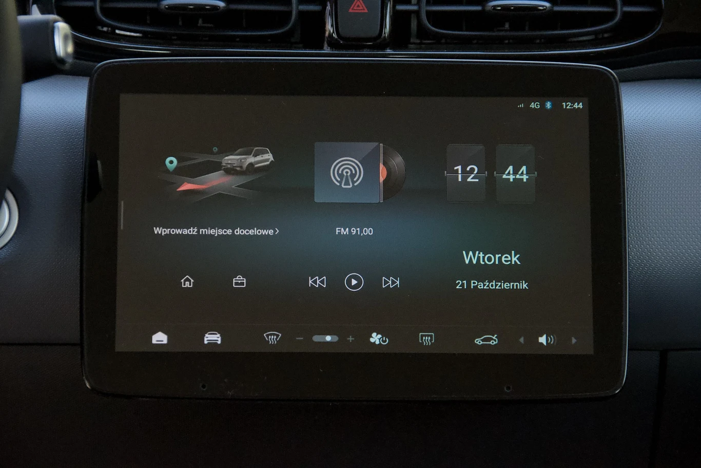 Duży ekran dotykowy systemu multimedialnego w samochodzie wyświetla informacje o nawigacji, radiu, godzinie oraz dacie. Elementy sterujące klimatyzacją i wentylacją widoczne w górnej części zdjęcia.