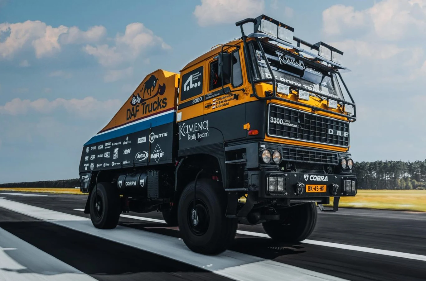 Ciężarówka DAF 3300 w sportowych barwach z licznymi naklejkami sponsorów, ustawiona na asfalcie, w tle widoczne rozległe pole i las pod błękitnym niebem.