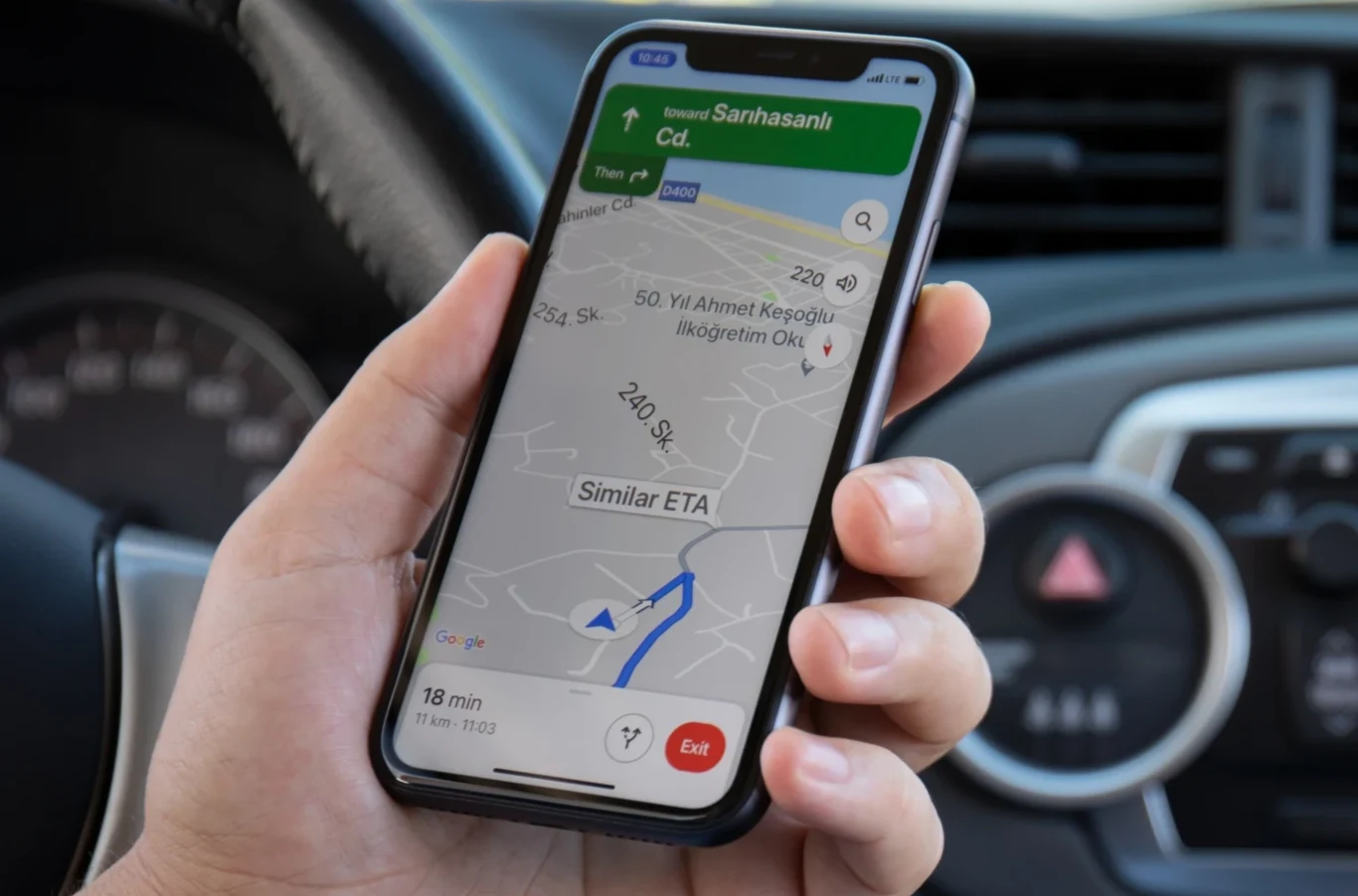 Jak obniżyć średnie spalanie samochodu przy użyciu nawigacji Google Maps? Dłoń trzymająca smartfon z uruchomioną aplikacją nawigacji GPS, w tle kokpit samochodu z widoczną kierownicą i deską rozdzielczą.