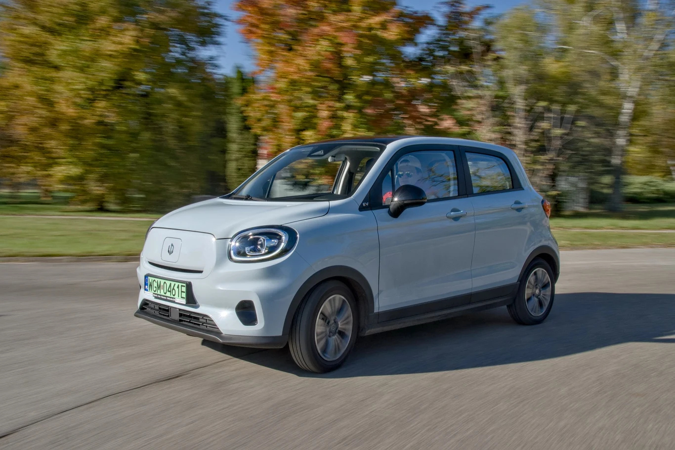 Mały, srebrny samochód elektryczny jedzie ulicą, otoczony drzewami w jesiennych kolorach. Dynamiczny kadr podkreśla ruch pojazdu, a zielona rejestracja wskazuje na ekologiczny charakter auta.