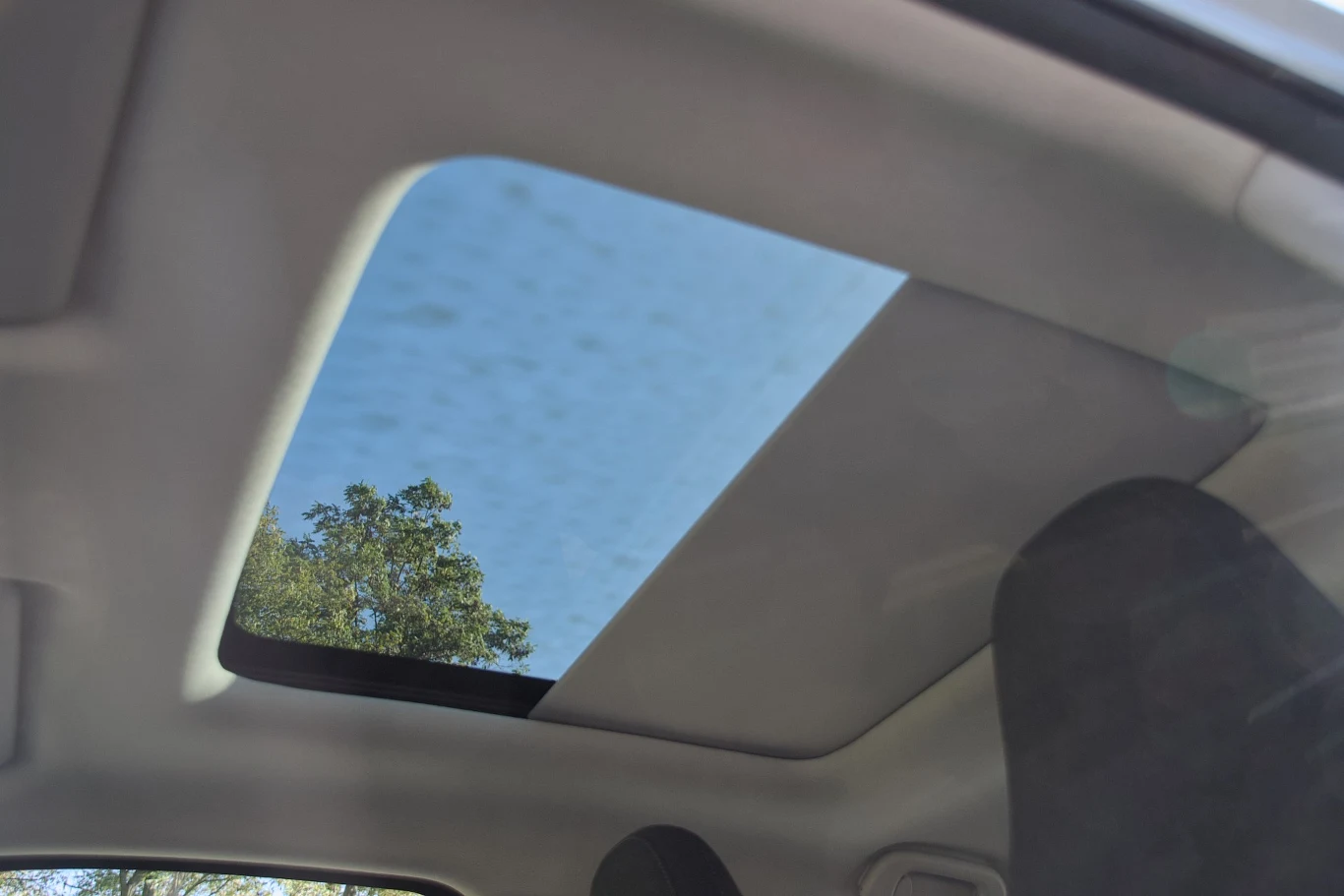 Szyberdach w samochodzie otwarty na tle nieba z delikatnie widoczną koroną drzewa. Wnętrze auta z częściowo widocznymi fotelami.