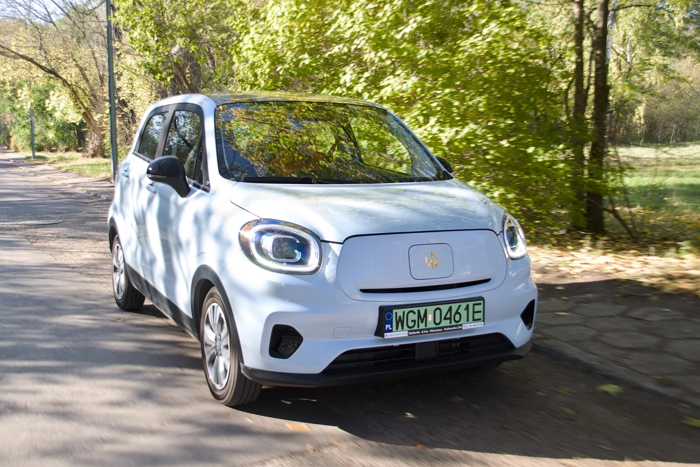 Mały biały samochód elektryczny poruszający się po asfaltowej drodze otoczonej zielonymi drzewami i krzewami w słoneczny dzień, z widoczną zieloną rejestracją.