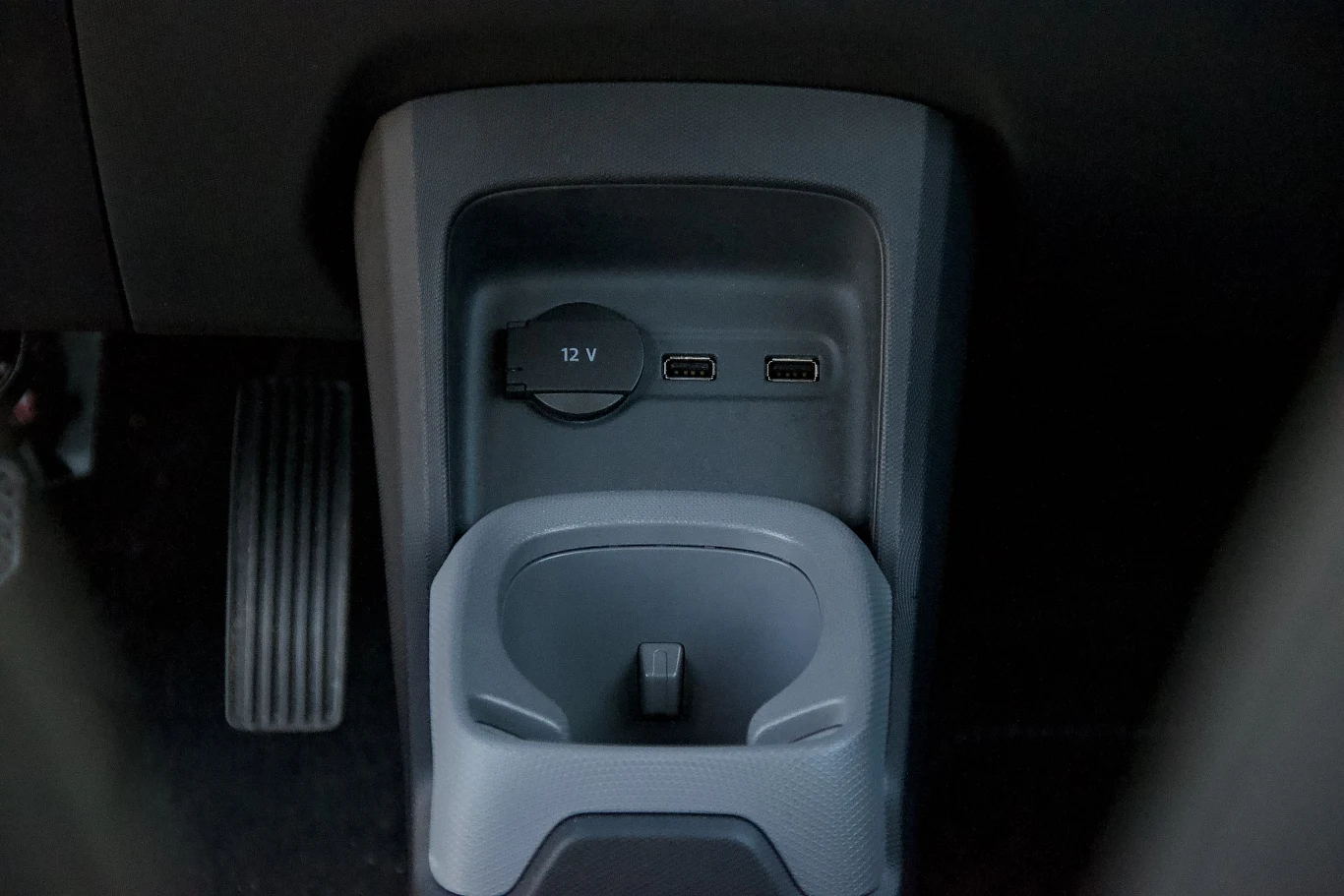 Konsola środkowa samochodu z gniazdem 12V, dwoma portami USB oraz uchwytem na napoje. Widoczny pedał przyspieszenia po lewej stronie.