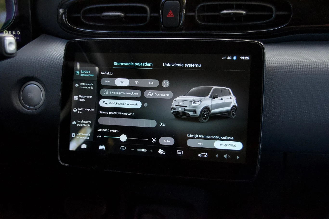 Dotykowy ekran komputera pokładowego samochodu wyświetla menu ustawień pojazdu, widoczna jest grafika samochodu, suwak jasności ekranu oraz opcje zarządzania światłami i alarmem. Wokół wyświetlacza znajdują się elementy wnętrza auta.