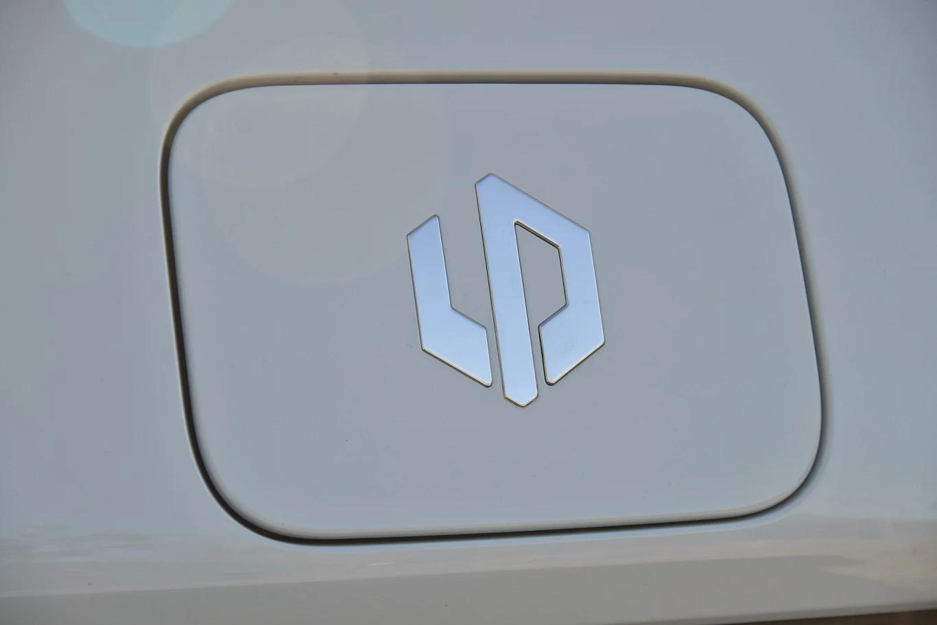 Chromowane logo o geometrycznym kształcie umieszczone na klapce wlewu paliwa białego samochodu.