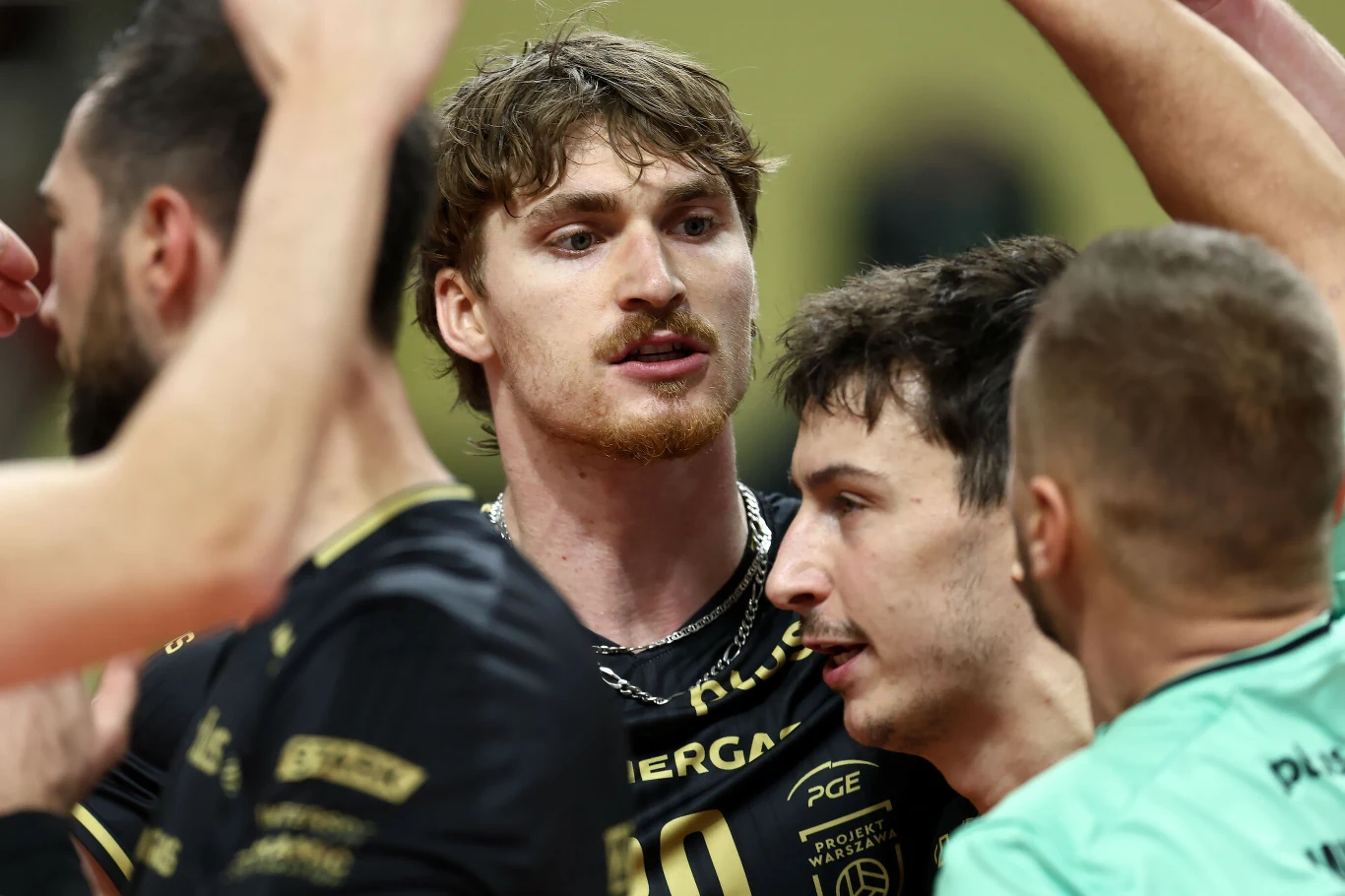 Grupa siatkarzy ubranych w sportowe stroje podczas intensywnej wymiany na boisku, jedna z postaci znajduje się centralnie w kadrze, wyrażając emocje po zakończonej akcji.