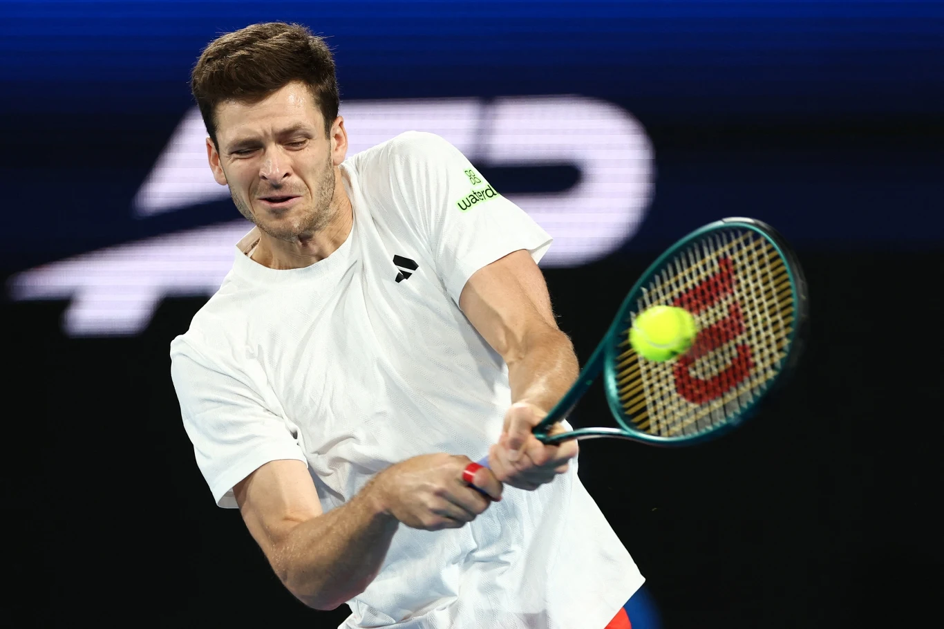 Hubert Hurkacz Tenisista wykonujący uderzenie backhandem podczas meczu, skupiony na lecącej piłce, ubrany w biały strój sportowy, na tle niebieskiego kortu.