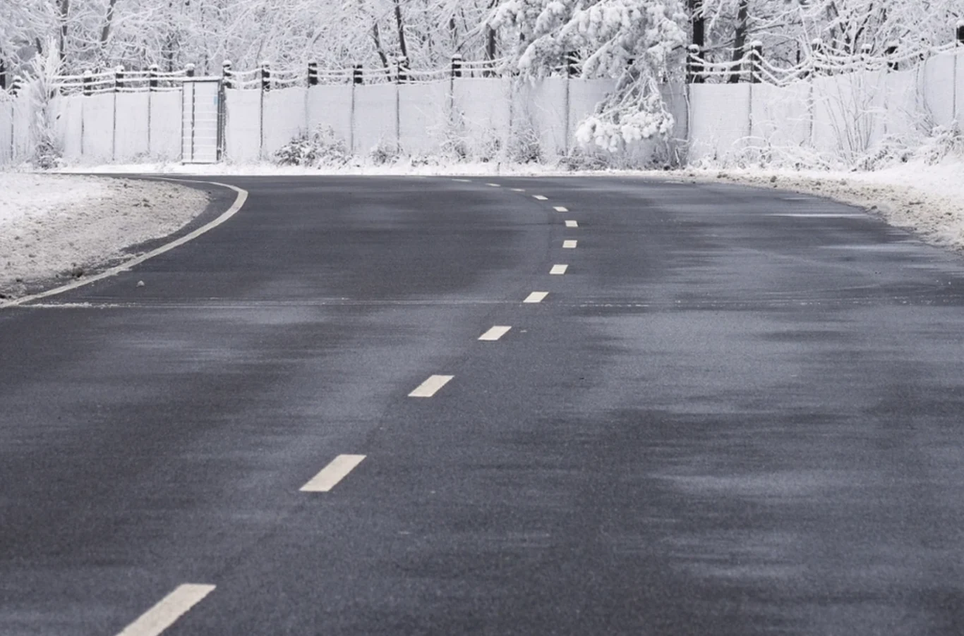 Tzw. czarny lód, gołoledź to zjawisko, które występuje na drogach od jesieni do wiosny. Asfaltowa droga prowadząca łagodnym łukiem przez krajobraz pokryty śniegiem, otoczona drzewami i ogrodzeniem, gałęzie i ziemia zasypane białym puchem, zimowa sceneria.