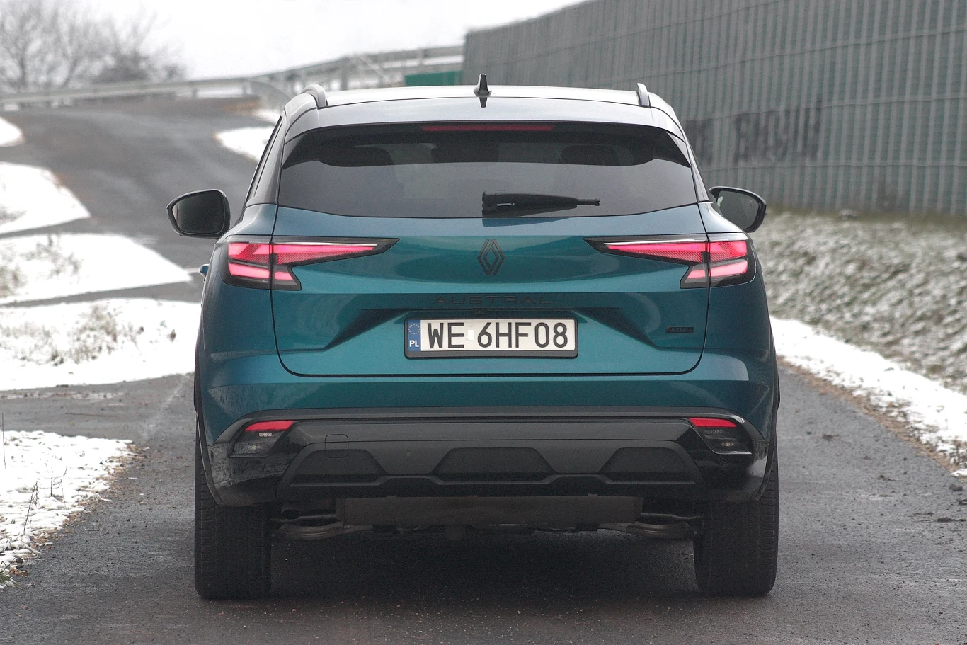 Zielony SUV marki Renault stoi na ośnieżonej drodze, otoczony cienką warstwą śniegu i metalowym płotem po prawej stronie. Widoczny jest głównie tył pojazdu, z podświetlonymi światłami i tablicą rejestracyjną.