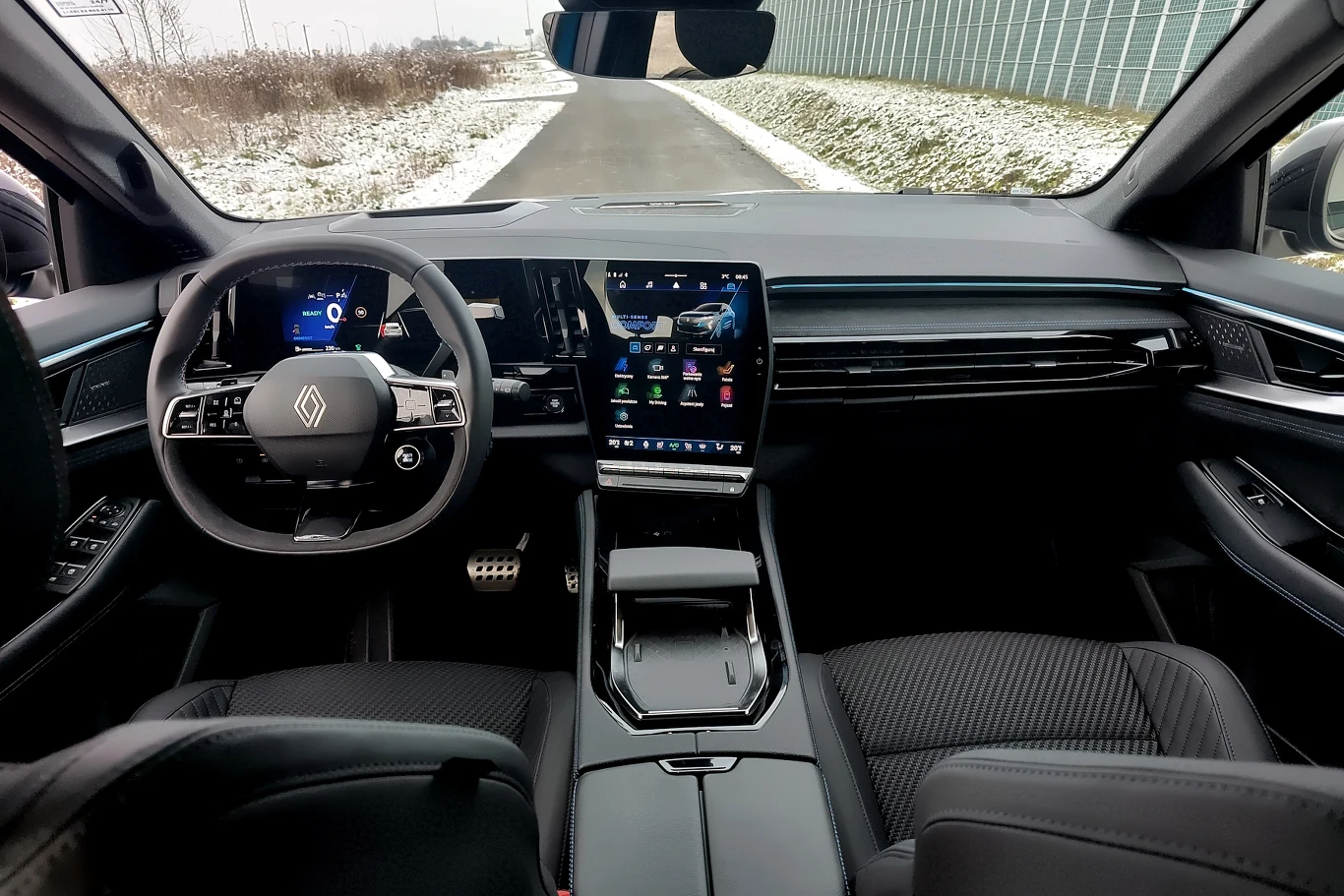 Nowoczesna deska rozdzielcza samochodu z dużym ekranem dotykowym na konsoli środkowej, cyfrowymi zegarami, kierownicą z logo producenta, wygodnymi fotelami i zimowym krajobrazem widocznym przez przednią szybę.