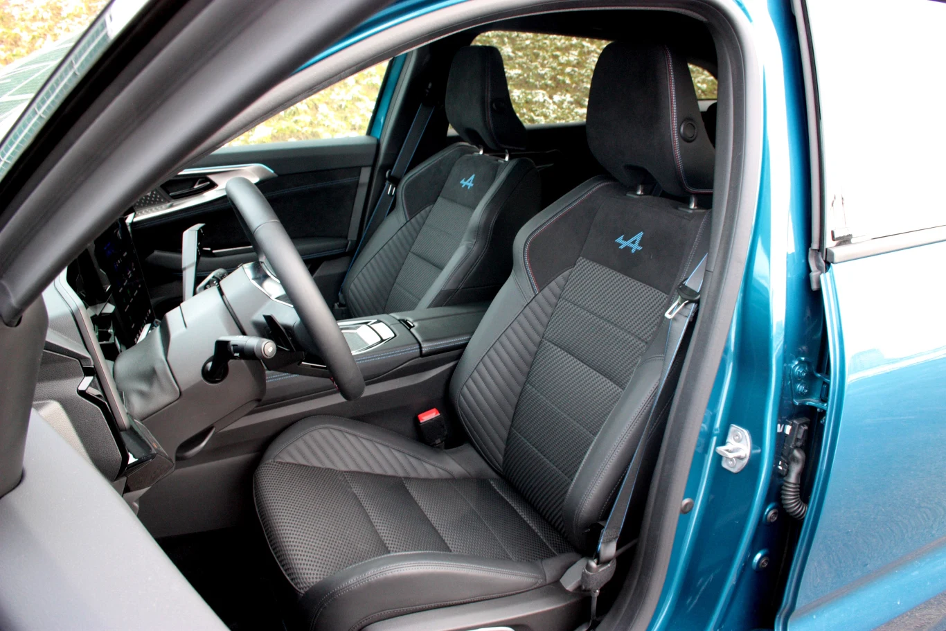 Wnętrze nowoczesnego samochodu z widocznymi przednimi fotelami z czarną tapicerką i logo Alpine, elegancka kierownica oraz dobrze zaprojektowana deska rozdzielcza, przeszklenie drzwi i fragment zewnętrznej karoserii w kolorze niebieskim.