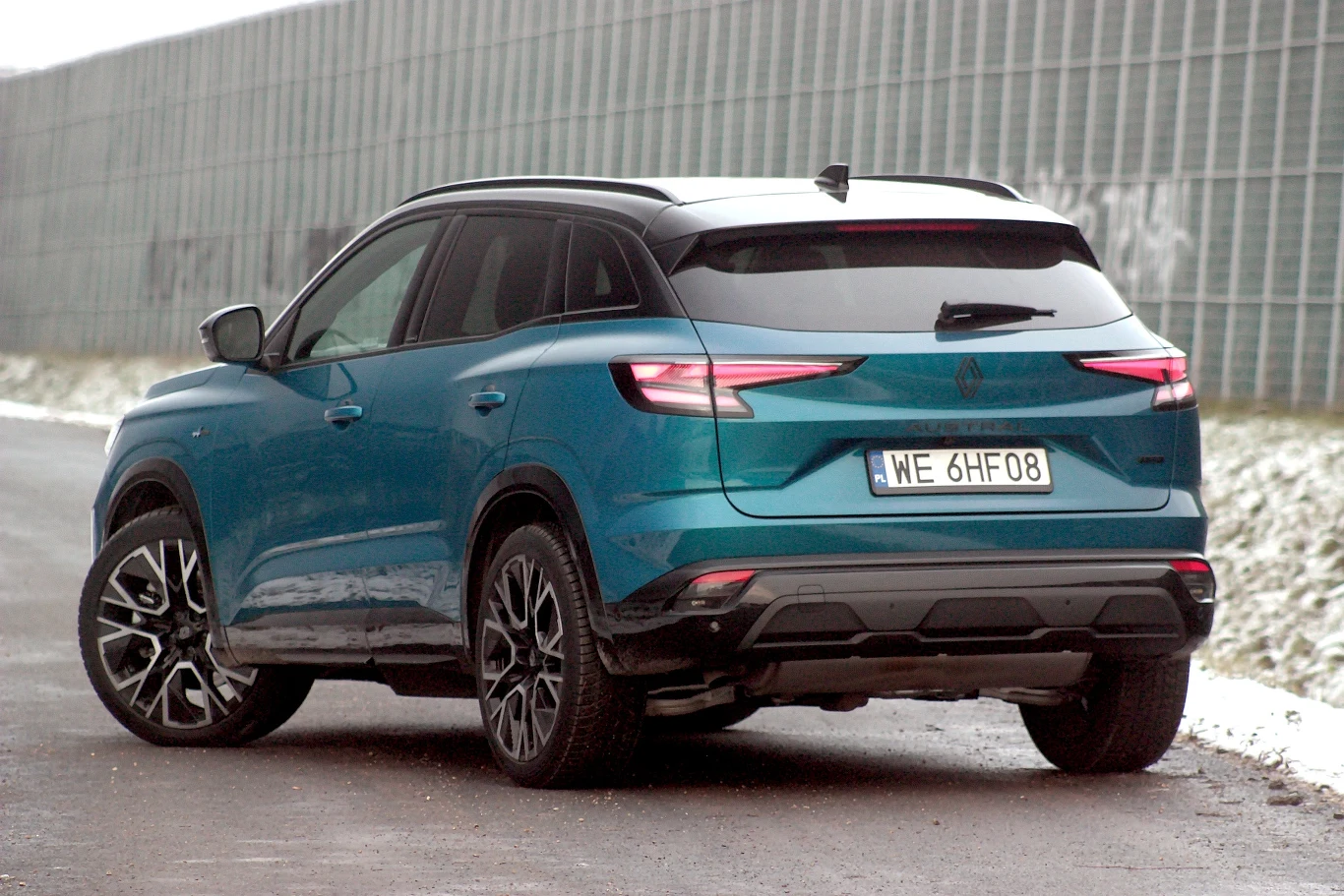 Nowoczesny, niebieski SUV zaparkowany przy drodze asfaltowej, z widoczną tylną tablicą rejestracyjną i masywnymi felgami, w tle szary ekran akustyczny i lekko ośnieżone pobocze.