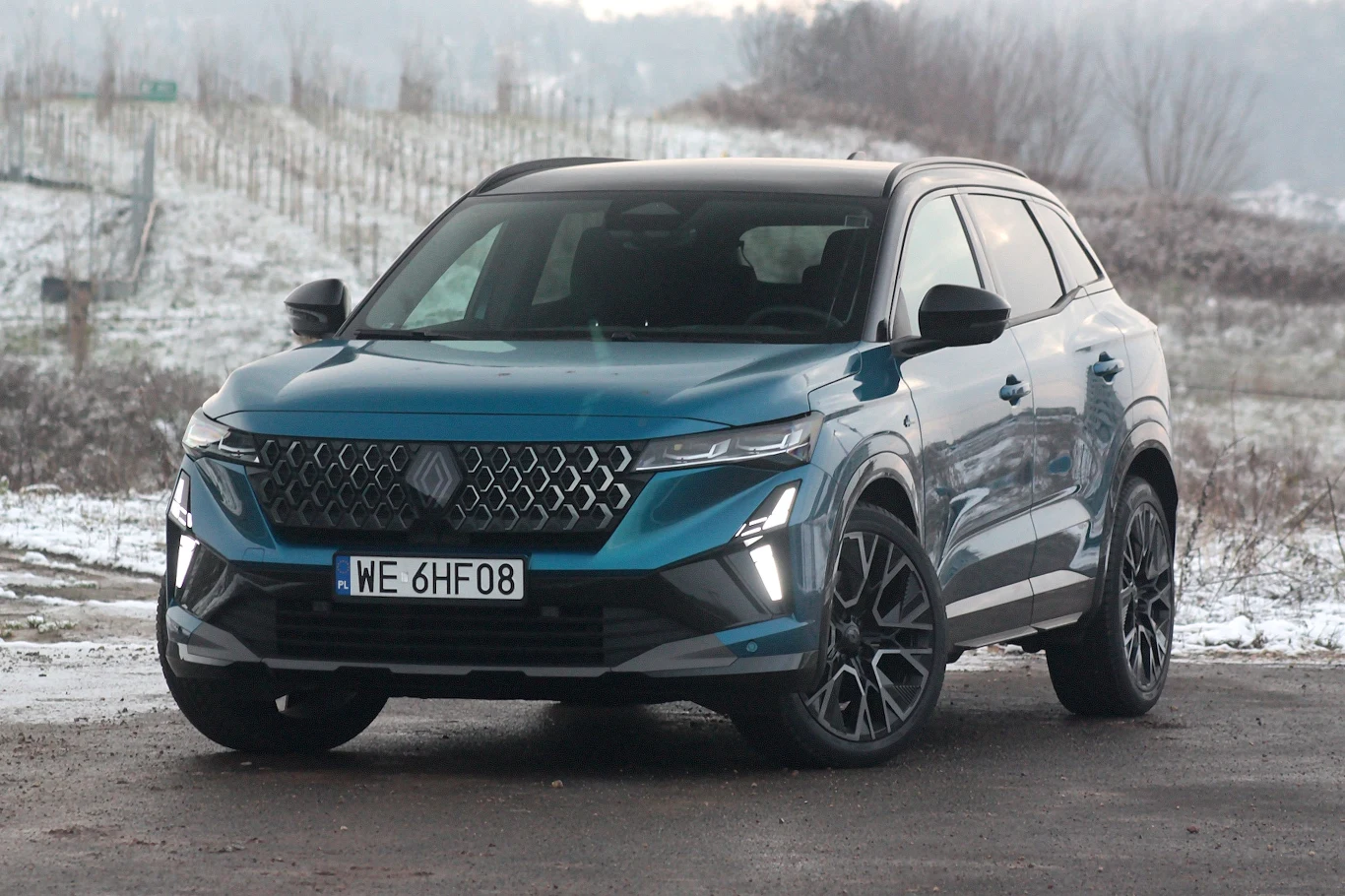 Nowoczesny, niebieski SUV marki Renault Austral zaparkowany na tle zimowego, lekko ośnieżonego krajobrazu. Światła dzienne LED o charakterystycznym kształcie, duże felgi oraz wyraźnie widoczna tablica rejestracyjna. Auto pokazane od przodu pod lekkim k...