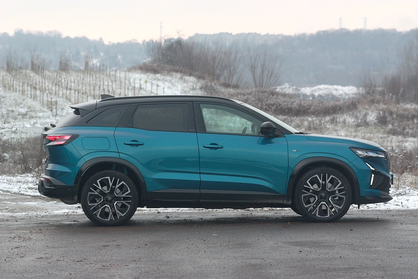 Nowoczesny, niebieski SUV zaparkowany na asfaltowym podłożu, na tle zimowego krajobrazu z zaśnieżonymi pagórkami i suchymi krzewami w oddali.