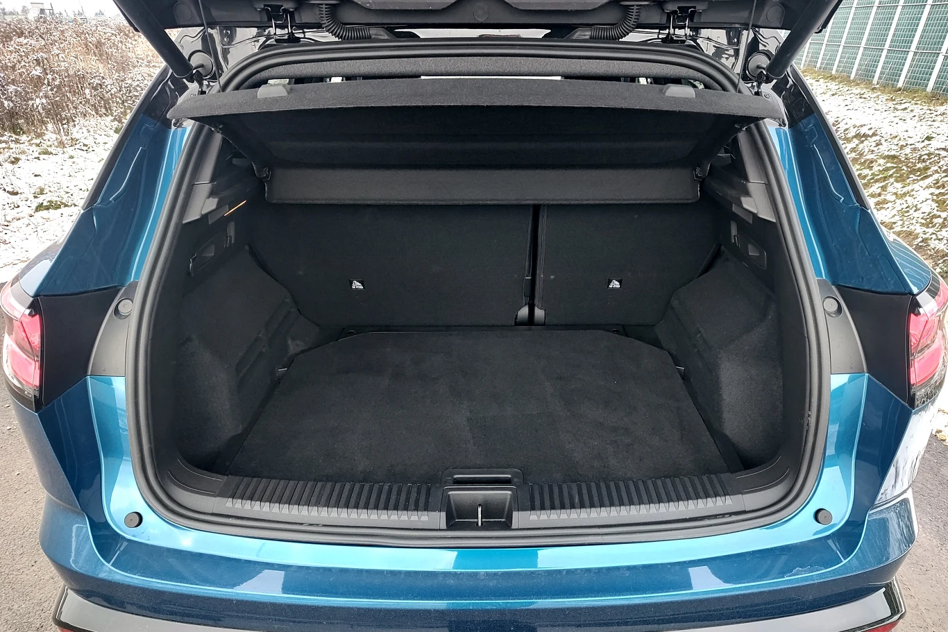 Otwarty bagażnik niebieskiego samochodu osobowego, widoczna przestrzeń ładunkowa z czystą tapicerką oraz automatycznie podnoszoną klapą. W tle fragment parkingu i ogrodzenie.