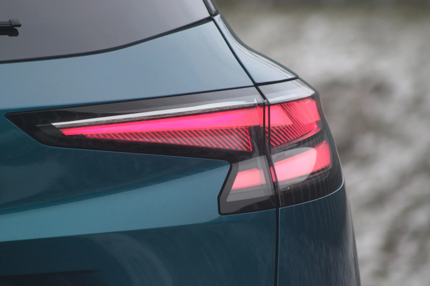 Tylna lampa nowoczesnego samochodu o aerodynamicznym kształcie, podkreślona świecącym, czerwonym światłem, zarysowana na tle rozmytego krajobrazu.
