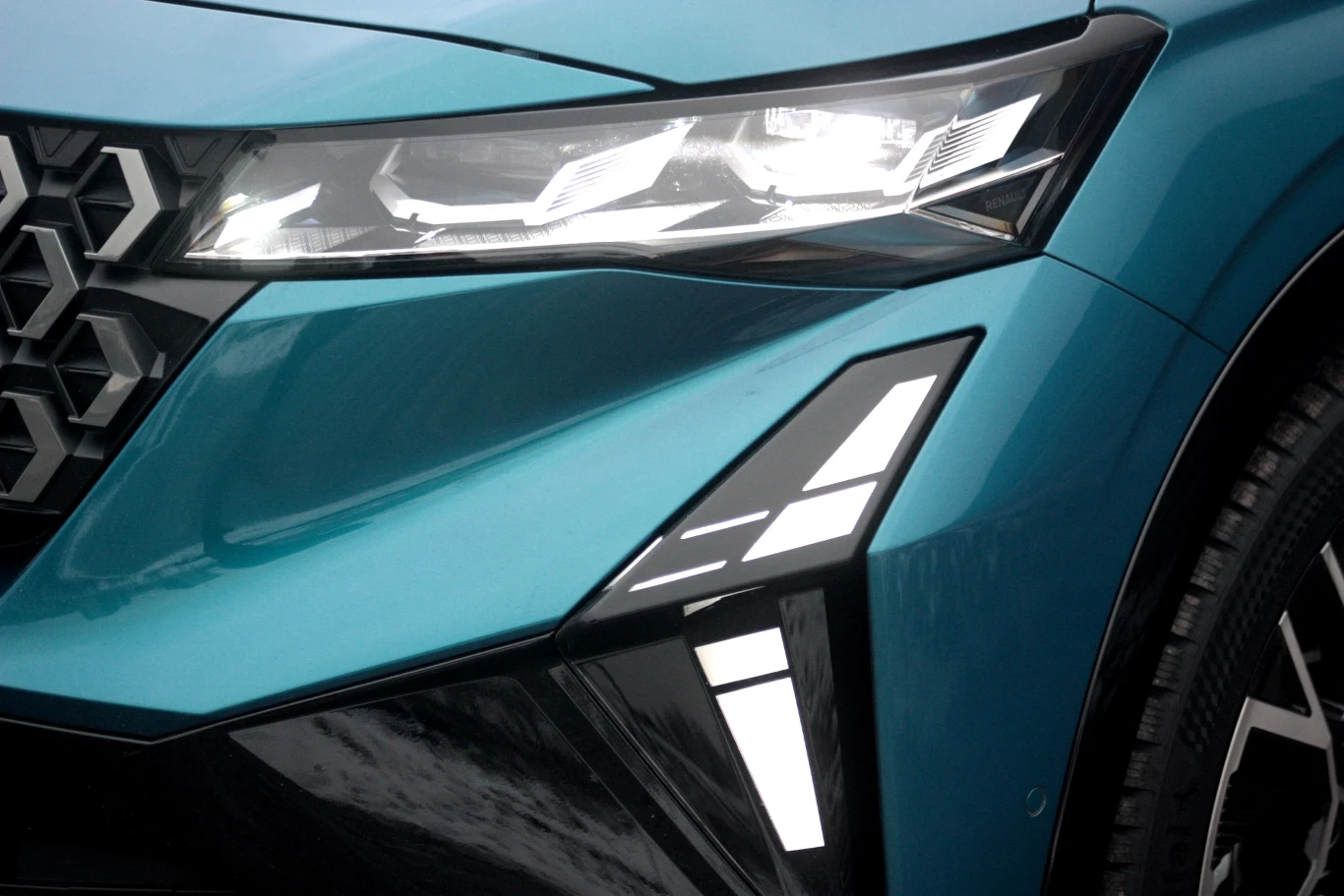 Przedni reflektor nowoczesnego samochodu z dynamicznymi liniami oraz efektownymi, ostrymi kształtami świateł LED, lakier w kolorze turkusowym, fragmenty kratki oraz opony widoczne w tle.
