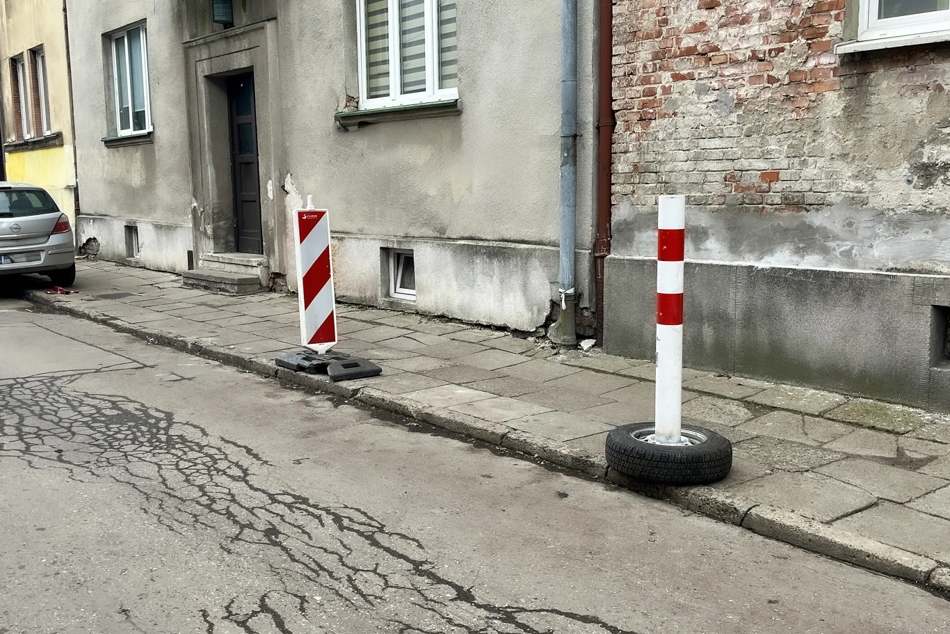 Blokujesz miejsce parkingowe? Prosisz się o mandat Słupki zabezpieczające chodnik, jeden z nich umieszczony w oponie, widoczne przy krawędzi starego chodnika i jezdni z popękanym asfaltem, otoczone elewacjami kamienic.