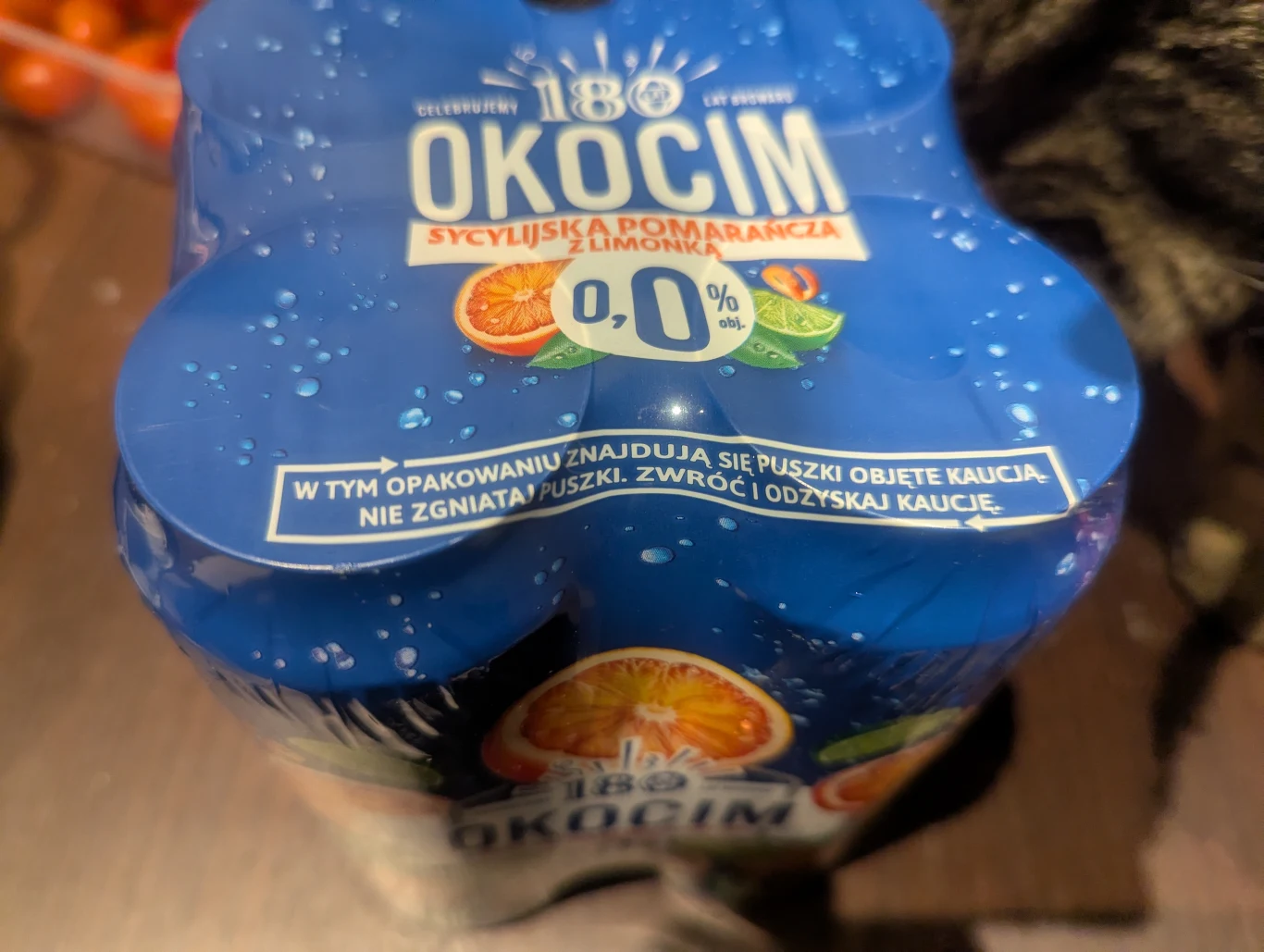 Zgrzewka puszek napoju bezalkoholowego Okocim o smaku sycylijskiej pomarańczy z limonką z wyraźnie zaznaczoną informacją o systemie kaucyjnym oraz zawartości 0,0% alkoholu.