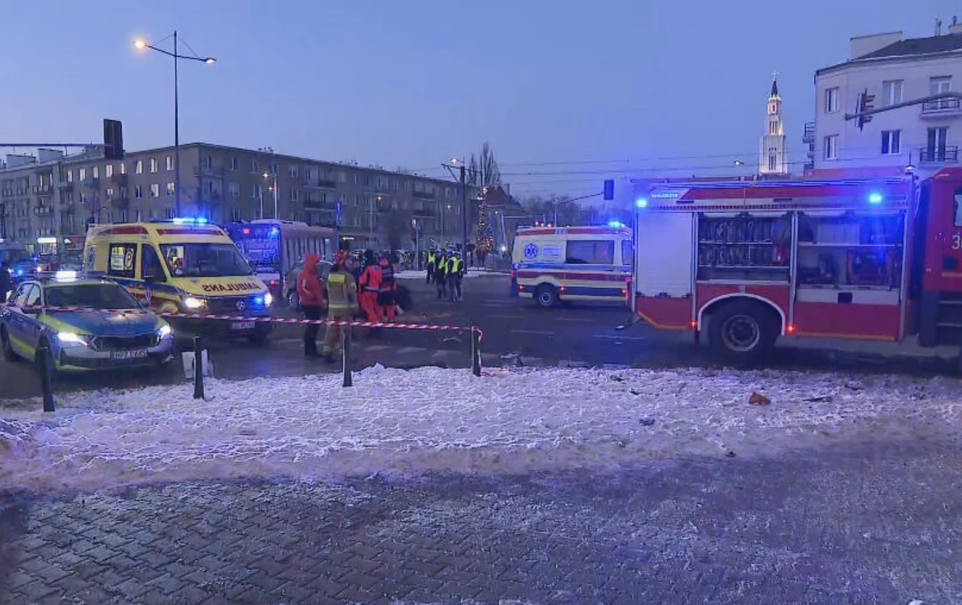Na skrzyżowaniu znajdują się pojazdy służb ratunkowych, w tym straż pożarna, policja i karetka pogotowia. Na ulicy stoi grupa ratowników, wokół widoczna jest taśma zabezpieczająca teren oraz ślady śniegu na chodniku. W tle widać zabudowania miejskie i ...