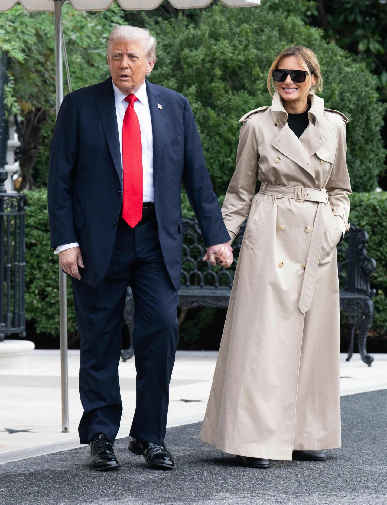 Mężczyzna w granatowym garniturze z czerwoną krawatą i kobieta w długim beżowym płaszczu trzymają się za ręce, spacerując na tle zieleni. Kobieta ma na sobie okulary przeciwsłoneczne i uśmiecha się. W tle widoczne krzewy oraz fragment ogrodzenia.