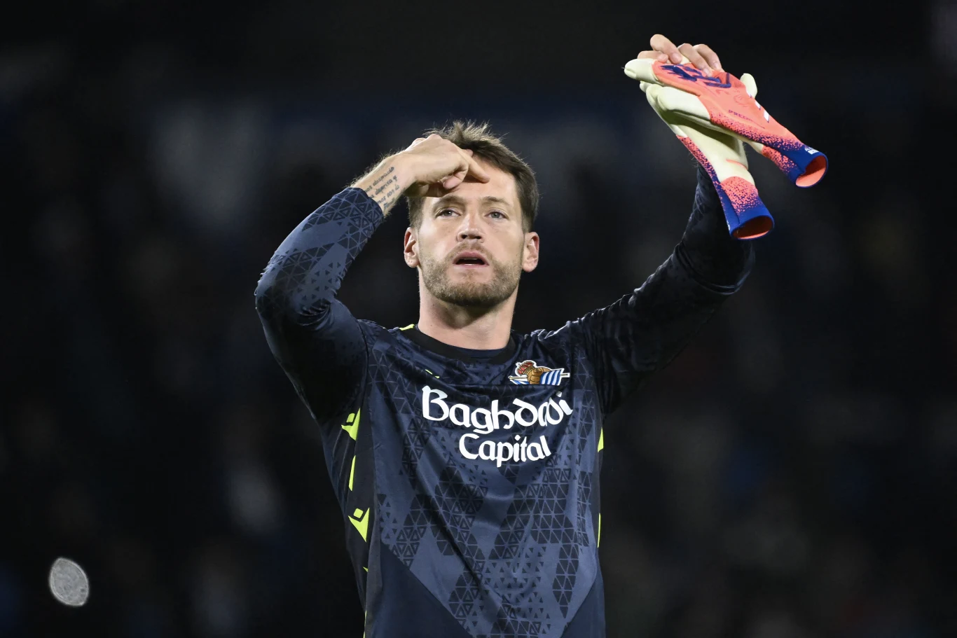 Alex Remiro Bramkarz w ciemnym stroju sportowym z logo sponsorów wykonuje gest, trzymając kolorowe rękawice bramkarskie w jednej ręce, na tle rozmytego stadionu.