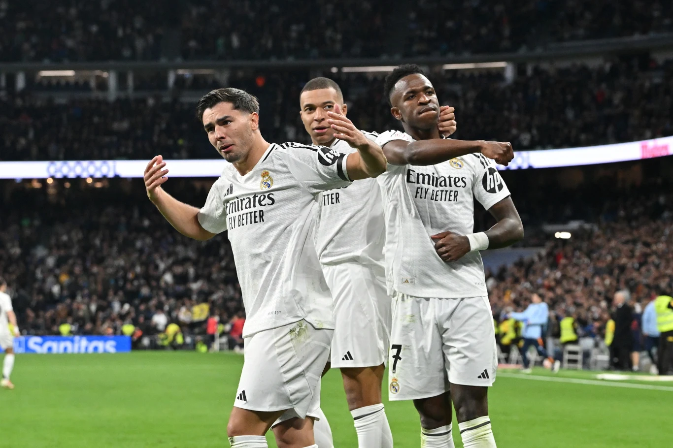 Piłkarze w białych strojach Realu Madryt celebrują zdobycie gola na tle pełnych trybun stadionu, trzeci z nich wykonuje charakterystyczny gest triumfu.