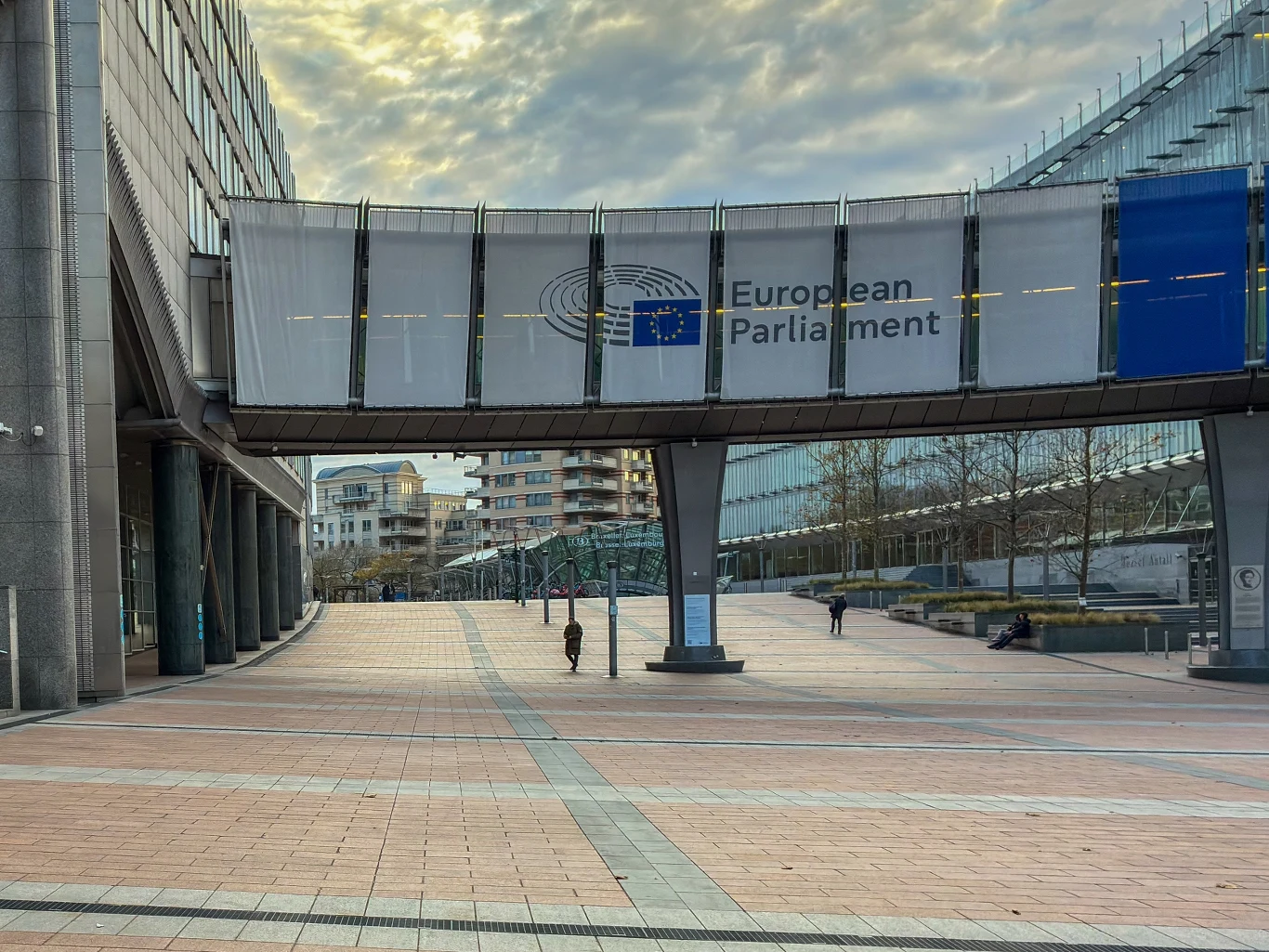 Parlament Europejski chce w trybie pilnym przegłosować pożyczkę dla Ukrainy Most łączący nowoczesne budynki Parlamentu Europejskiego z widocznym napisem, na pierwszym planie szeroka promenada, kilku spacerujących ludzi oraz przeszklone fasady biurowców w tle.