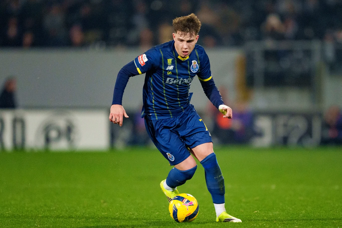 Młody piłkarz w granatowym stroju drużyny Porto prowadzi piłkę podczas meczu na murawie, skupiony na grze, w tle widoczne rozmyte trybuny ze słabo rozpoznawalnymi kibicami.