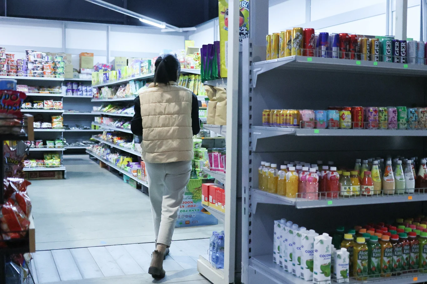 System kaucyjny. Producenci znaleźli sposób na omijanie przepisów (zdj. ilustracyne) Kobieta w jasnej kamizelce przechadza się alejką w dobrze oświetlonym sklepie spożywczym, na półkach widoczne są różnorodne napoje, przekąski i produkty spożywcze.