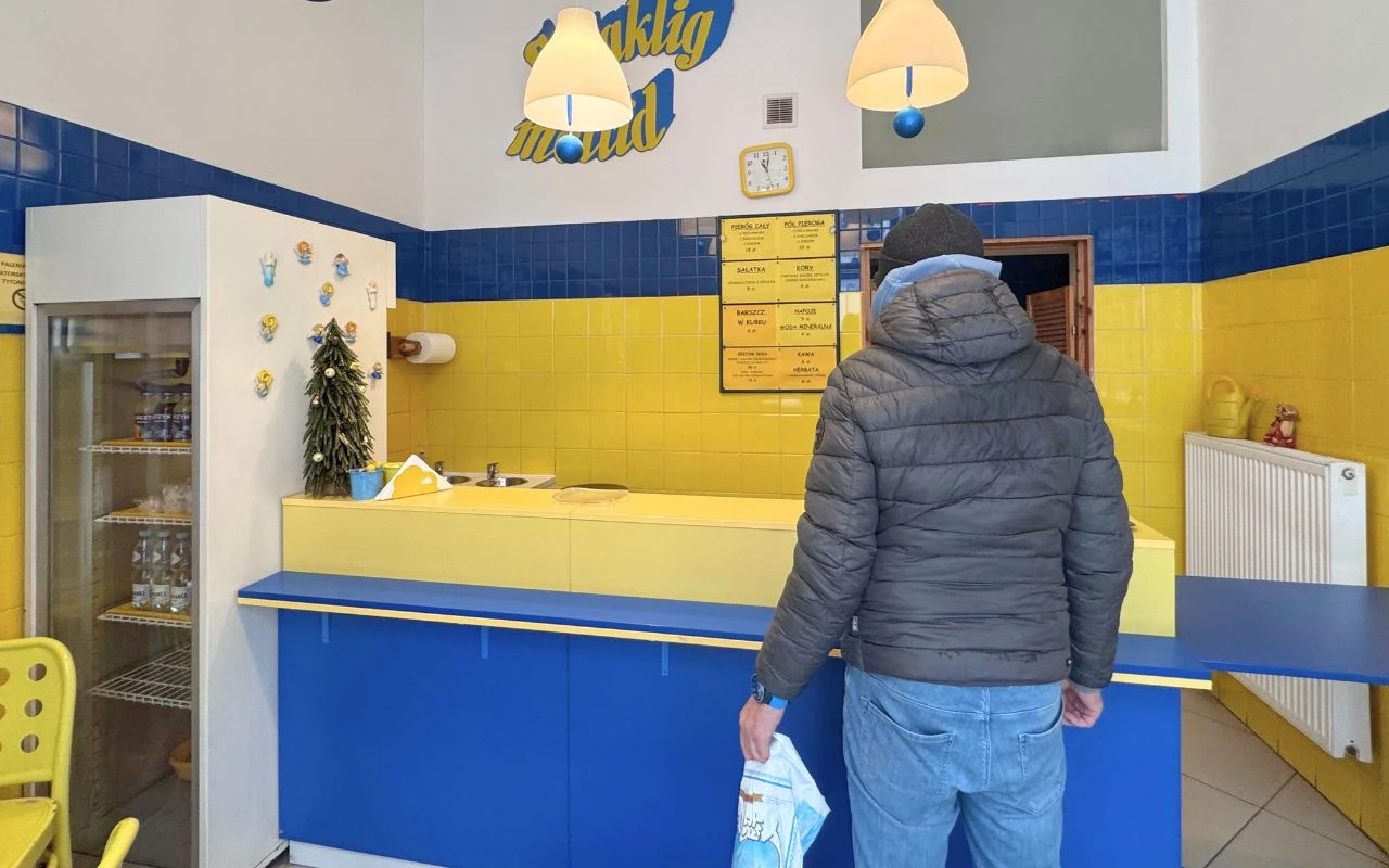 Mężczyzna w kurtce i czapce stoi przy ladzie niewielkiego lokalu gastronomicznego utrzymanego w żółto-niebieskiej kolorystyce, z menu na ścianie i lodówką po lewej stronie; wnętrze minimalistyczne, dobrze oświetlone lampami sufitowymi.