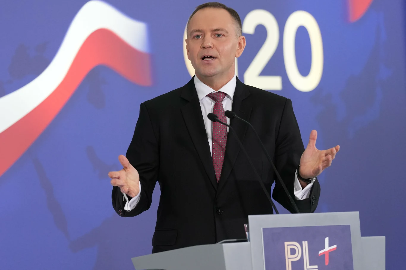 Mężczyzna w garniturze przemawia przy mównicy na tle flagi Polski i dużych cyfr 2020 oraz napisu PL+