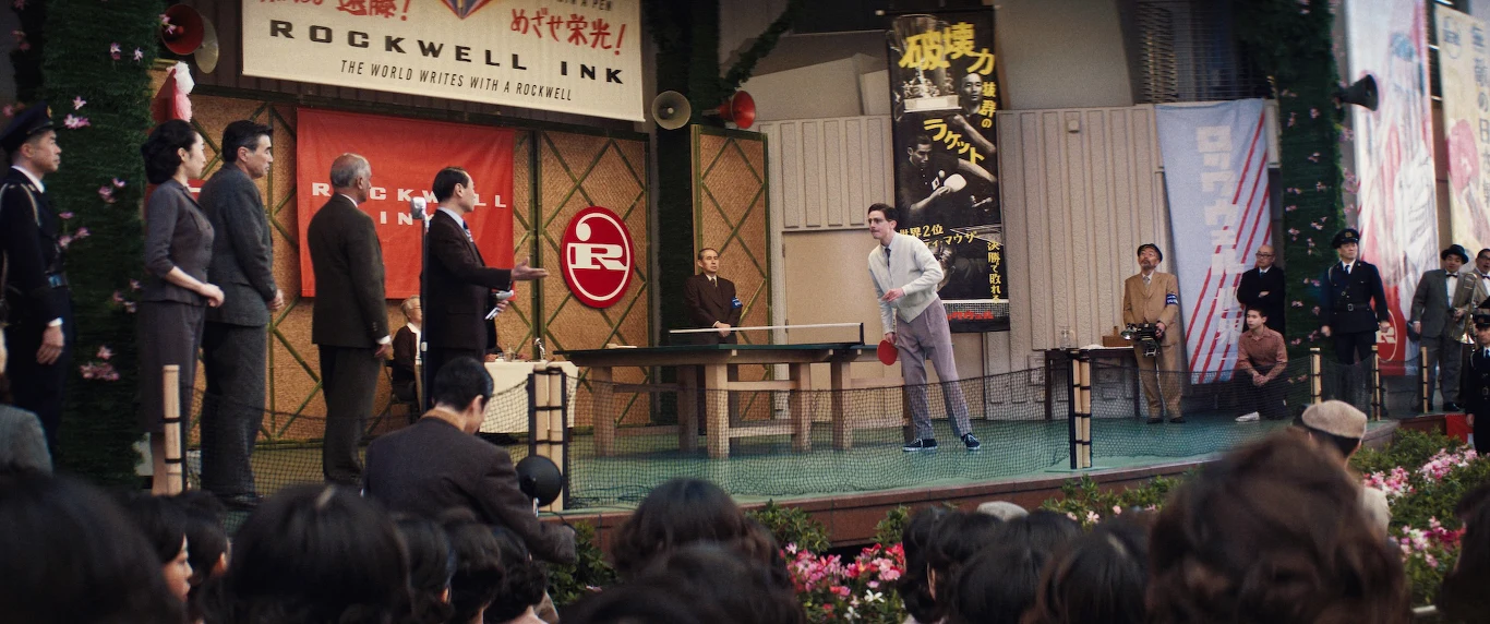 Mężczyzna grający w tenisa stołowego na scenie przed dużą publicznością, po bokach widoczni obserwatorzy i sędziowie, w tle banery oraz dekoracje nawiązujące do sportu i azjatyckiej kultury.