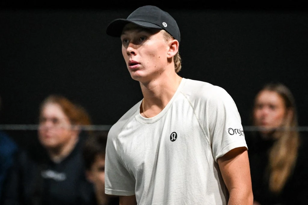 Młody mężczyzna w białej koszulce sportowej i czarnej czapce z daszkiem stoi w lekkim półprofile, w tle rozmyte sylwetki kilku osób obserwujących wydarzenie.