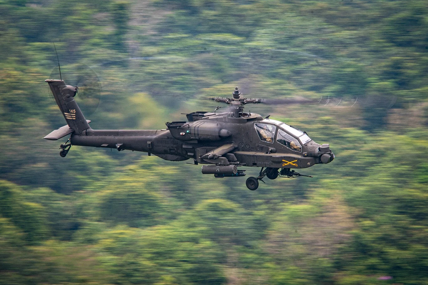 Wojskowy śmigłowiec bojowy lecący nad zielonym, zalesionym terenem z dobrym widokiem na konstrukcję maszyny i umieszczone na niej oznaczenia.