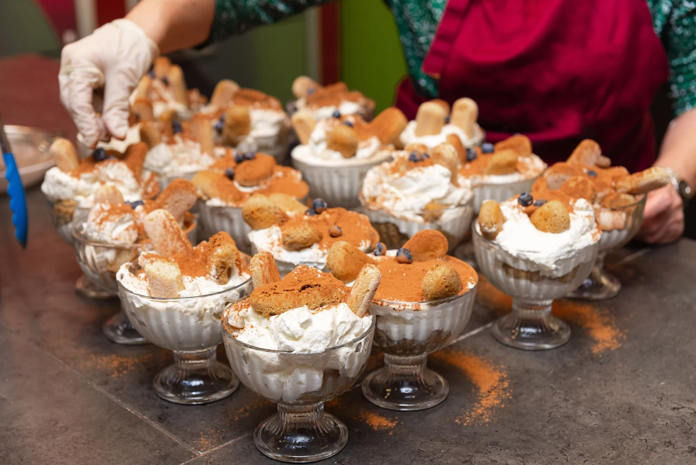 Wiele pucharków z deserem tiramisu, wypełnionych kremem, biszkoptami, posypanych kakao i udekorowanych owocami, ustawionych na blacie. Osoba w fartuchu dekoruje desery, używając rękawiczek.