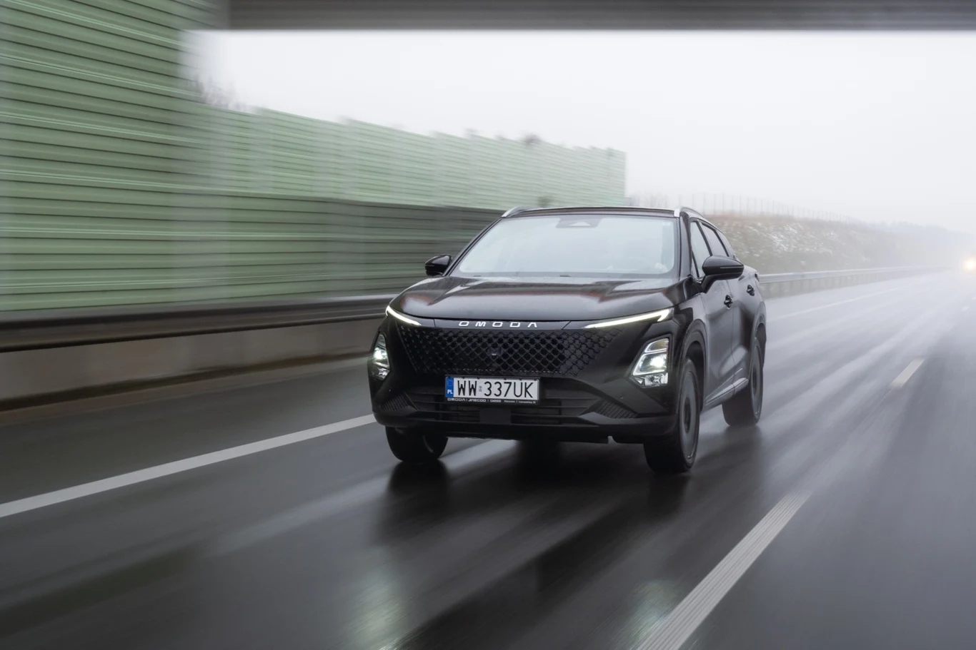 OMODA 5 Hybrid oferuje nie tylko atrakcyjne spalanie, ale także niezłe osiągi Nowoczesny samochód terenowy koloru czarnego jedzie po mokrej, szerokiej jezdni pod wiaduktem. W tle widoczne bariery dźwiękochłonne oraz rozmyte elementy krajobrazu, typowa sytuacja drogowa podczas pochmurnej pogody.