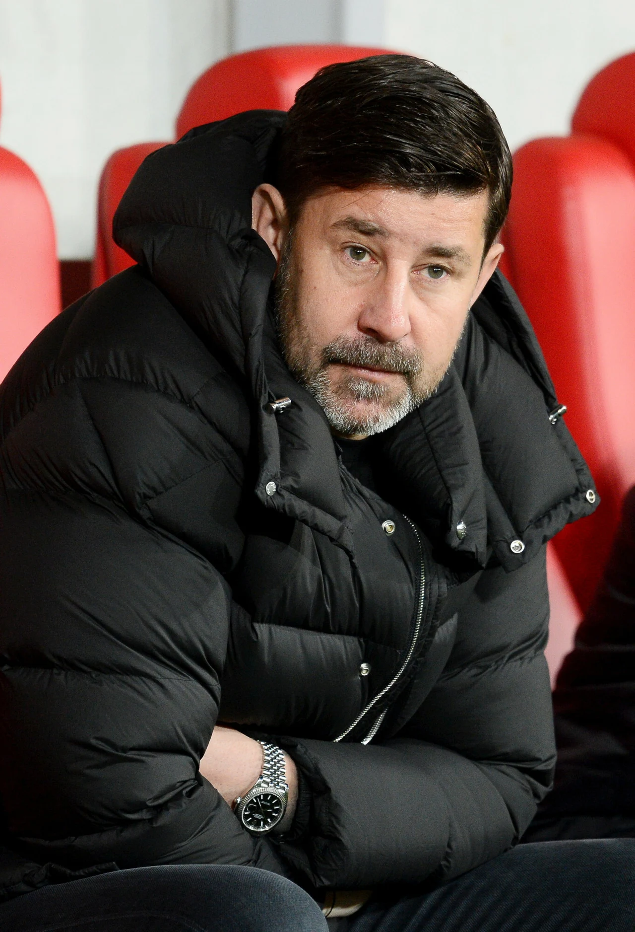 Mężczyzna z krótką brodą i ciemnymi włosami siedzący na czerwonym fotelu w puchowej, czarnej kurtce, z poważnym wyrazem twarzy i rękami założonymi na siebie.