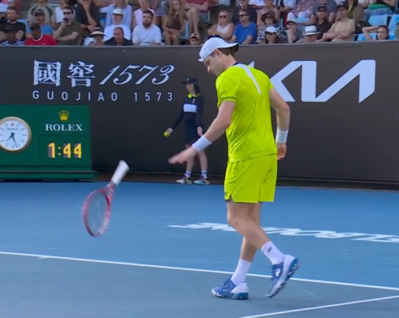 Tenisista w żółtym stroju na korcie podczas turnieju, trzymający rakietę odbijającą piłkę, tłum kibiców i sędzia w tle, elektroniczny zegar oraz reklamy na bandach.