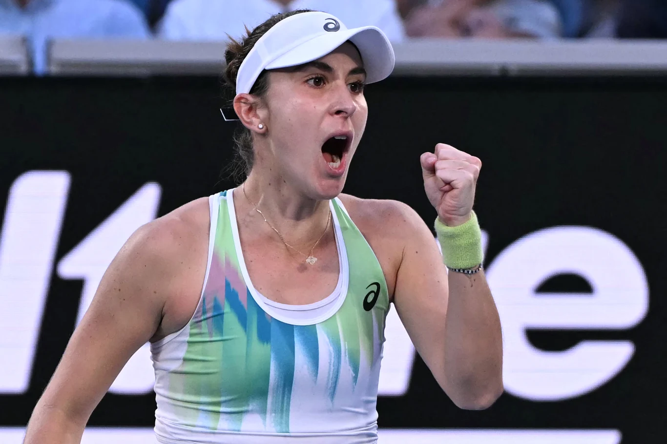 Tenisistka na korcie w sportowym stroju, z podniesioną zaciśniętą pięścią i wyrazem triumfu oraz skupienia na twarzy, w tle widoczne rozmazane elementy trybun.