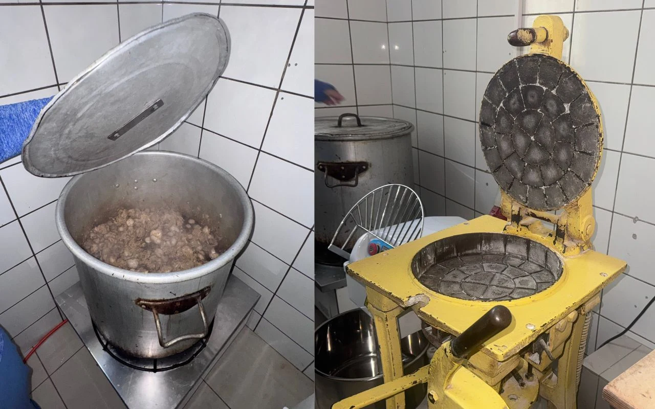 Duży metalowy garnek wypełniony gotującą się żywnością oraz żółta maszyna do wypieku wafli lub placków, ustawione w kafelkowanym pomieszczeniu kuchennym.