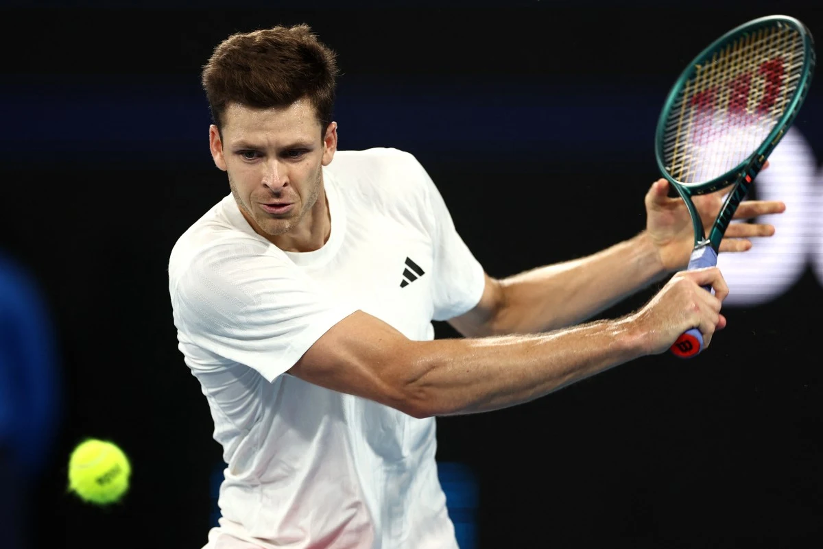 Sportowiec w białej koszulce wykonuje dynamiczne uderzenie rakietą tenisową, koncentrując się na odbiciu nadlatującej piłki tenisowej podczas meczu na korcie.