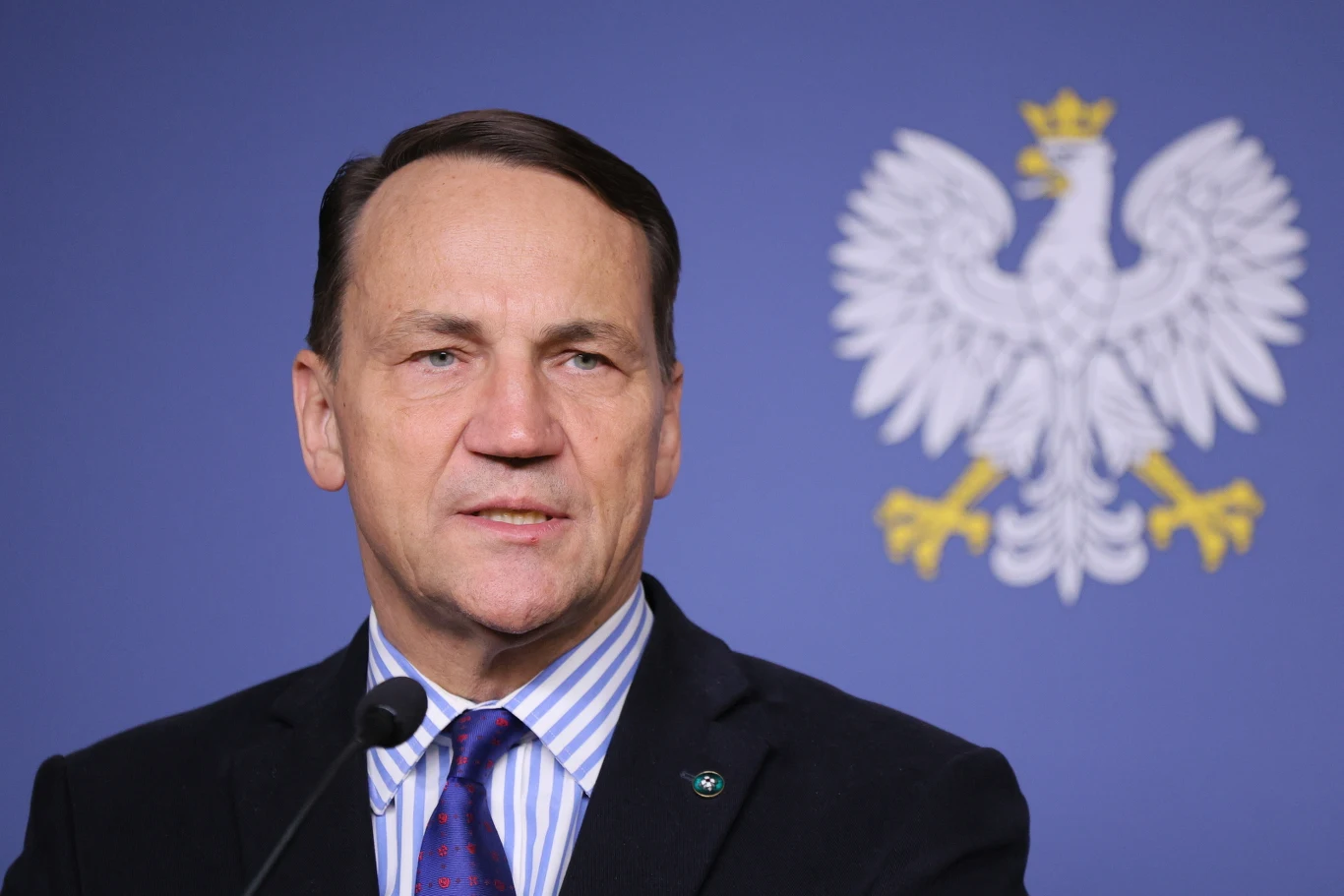 Szef MSZ Radosław Sikorski Mężczyzna w garniturze i krawacie przemawia przy mikrofonie na tle niebieskiej ściany z białym godłem Polski.