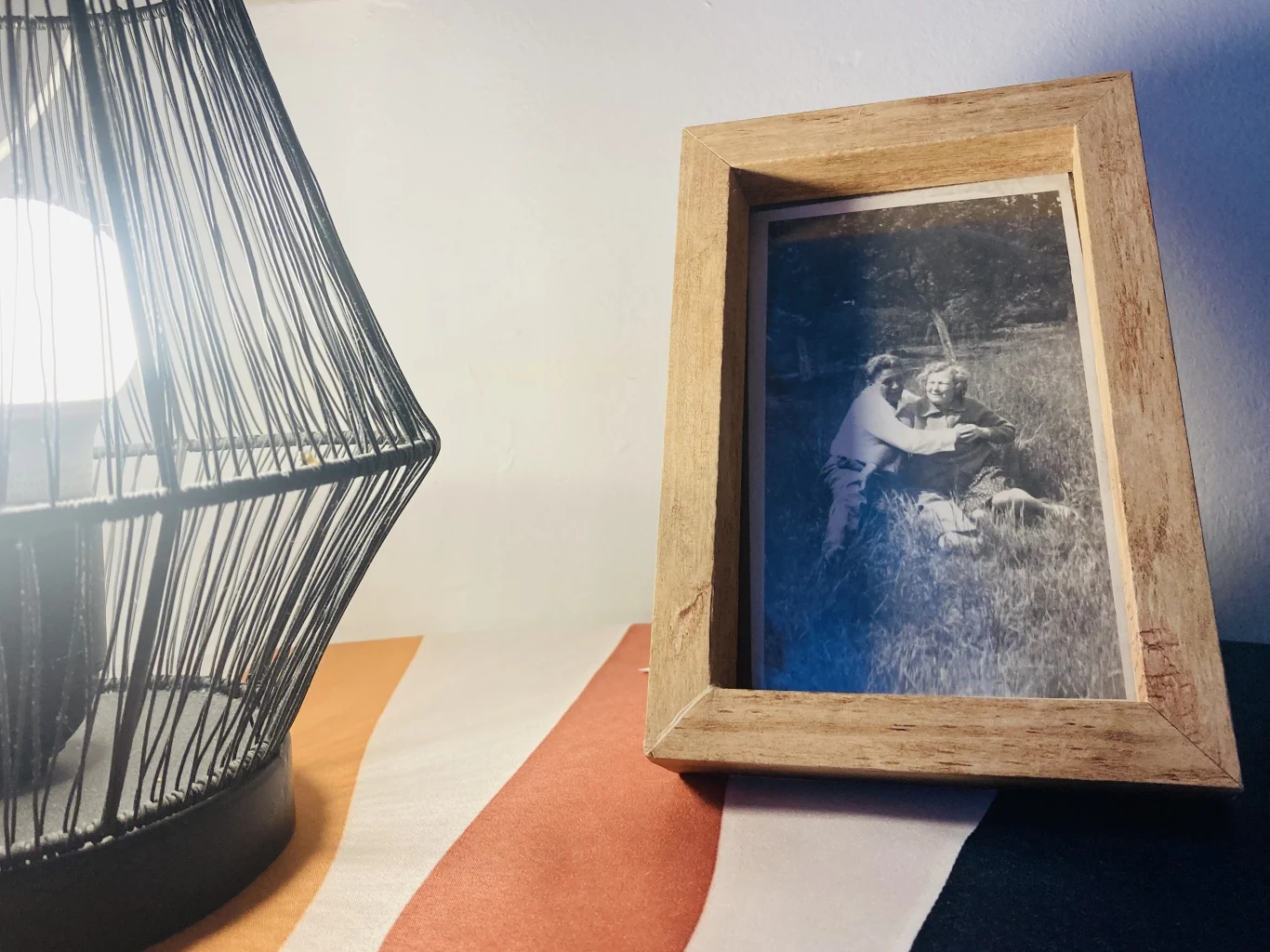 Drewniana ramka ze starym czarno-białym zdjęciem ustawiona na kolorowej tkaninie obok metalowej lampy emitującej ciepłe światło.