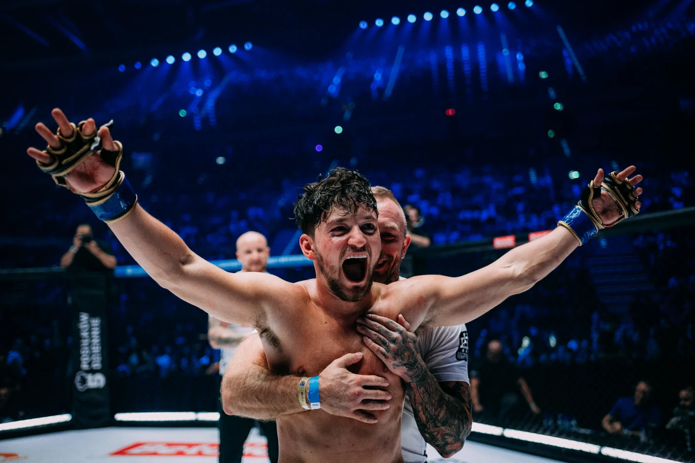 Zawodnik MMA z szeroko rozpostartymi ramionami i okrzykiem triumfu na twarzy, obejmowany przez trenera. W tle widoczne światła areny oraz publiczność obserwująca wydarzenie.