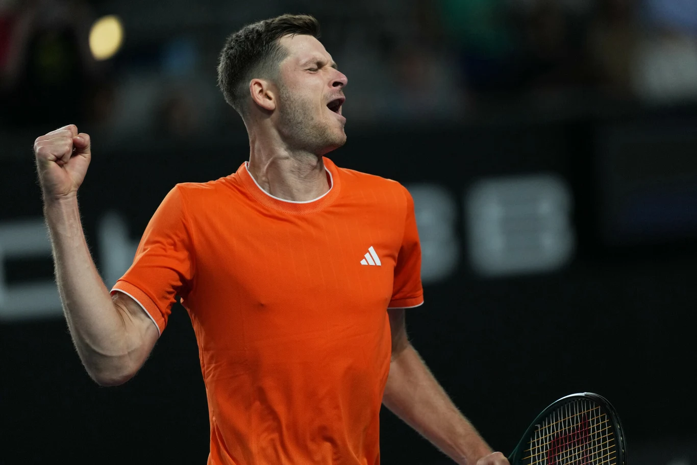 Mężczyzna w pomarańczowej koszulce sportowej z uniesioną pięścią w geście triumfu podczas meczu tenisowego, w ręce trzyma rakietę tenisową.