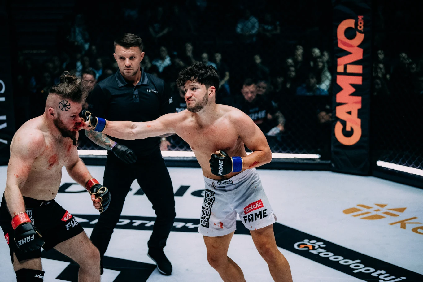 Dwóch zawodników walczy w oktagonie, jeden z nich zadaje cios przeciwnikowi prosto w twarz, a sędzia uważnie obserwuje przebieg pojedynku. Na podłodze i bandach widoczne są logotypy sponsorów.