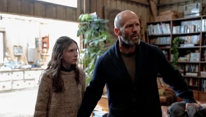 Mężczyzna w swetrze stoi obok młodej dziewczynki w jasnym ubraniu w jasnym, przytulnym pomieszczeniu z drewnianymi ścianami i regałami z książkami, w tle widać roślinę doniczkową i rozświetlone okno.