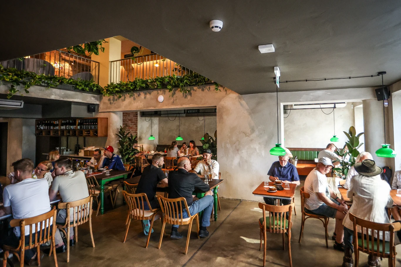 Ludzie siedzą przy stołach w restauracji o nowoczesnym wystroju, rozmawiając i spożywając posiłki, zielone lampy nad stołami, roślinność na antresoli oraz ciepłe, naturalne światło wnętrza podkreślają przyjazną atmosferę miejsca.